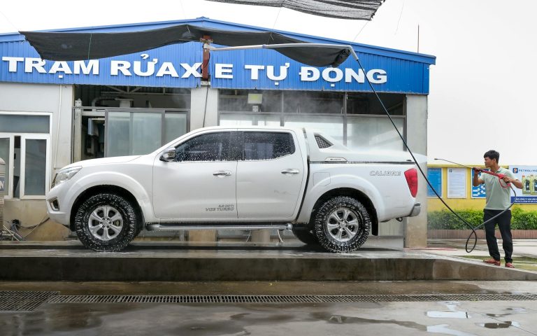 Adult using pressure washer to clean a white pickup truck at an outdoor car wash facility.